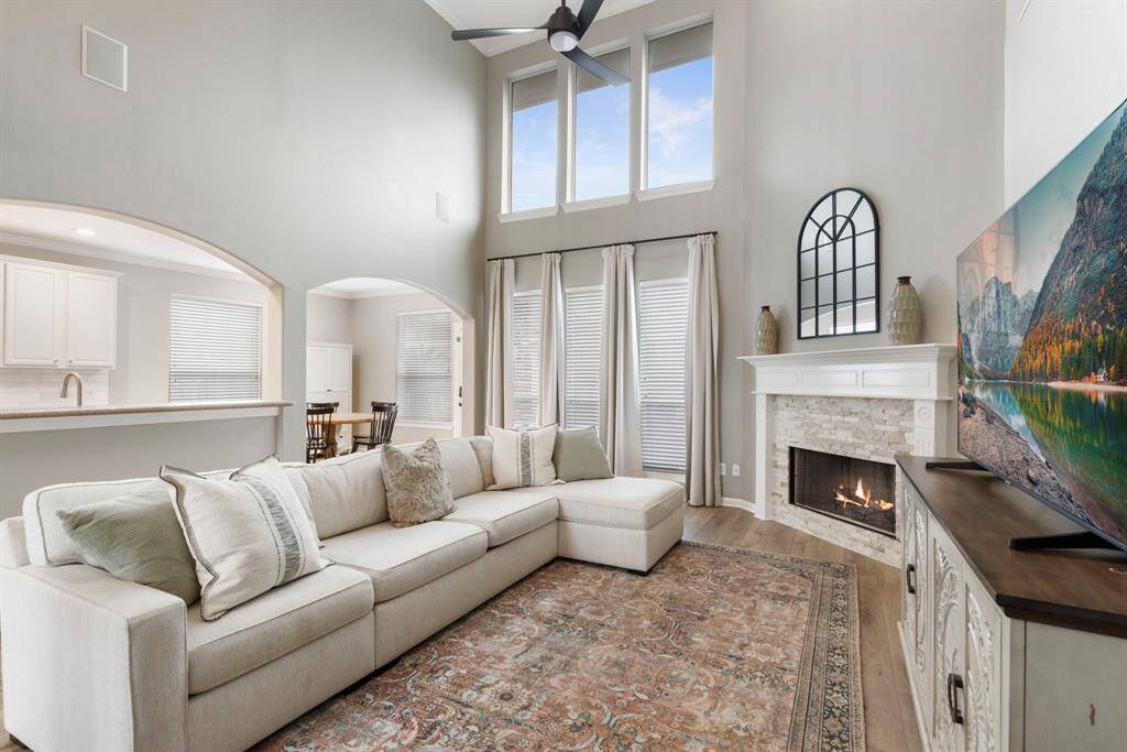 This spacious living room features high ceilings, large windows for natural light, a cozy fireplace, and an open layout connecting to the kitchen and dining area. It offers a comfortable, modern aesthetic with neutral tones and elegant decor.