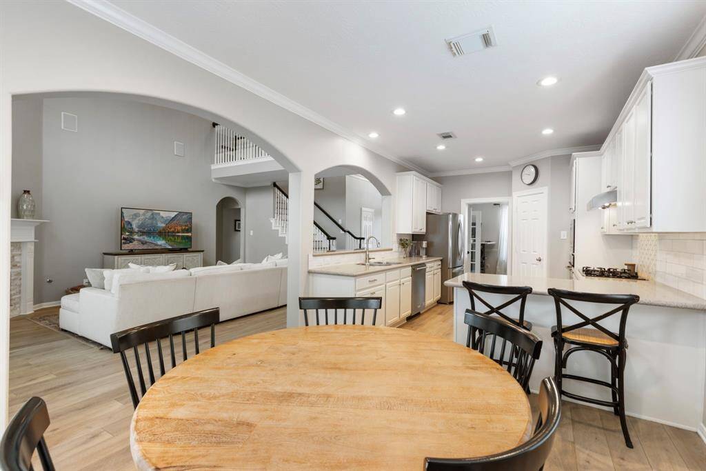 This photo showcases an open-concept kitchen and living area, featuring a cozy dining space. The modern kitchen has white cabinetry, stainless steel appliances, and a breakfast bar. The living room is spacious with ight wood flooring, and a view of a staircase leading to the upper level. The neutral color palette creates a bright and inviting atmosphere.