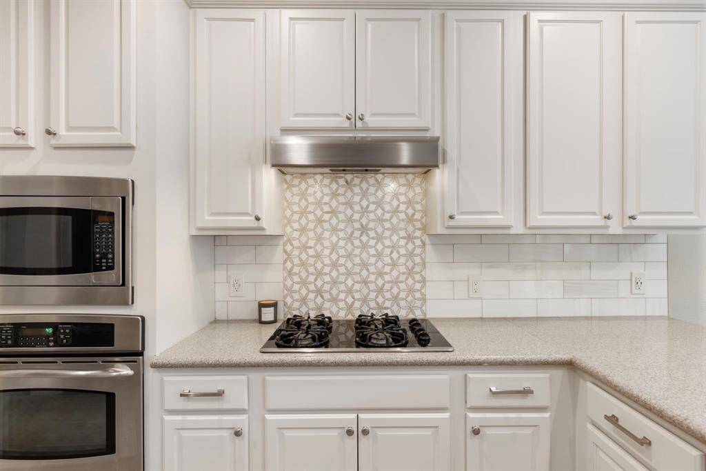 This kitchen features sleek stainless steel appliances, including a built-in oven and microwave. It has elegant white cabinetry, a stylish patterned backsplash above the stove, and a modern vent hood, creating a clean and contemporary look.