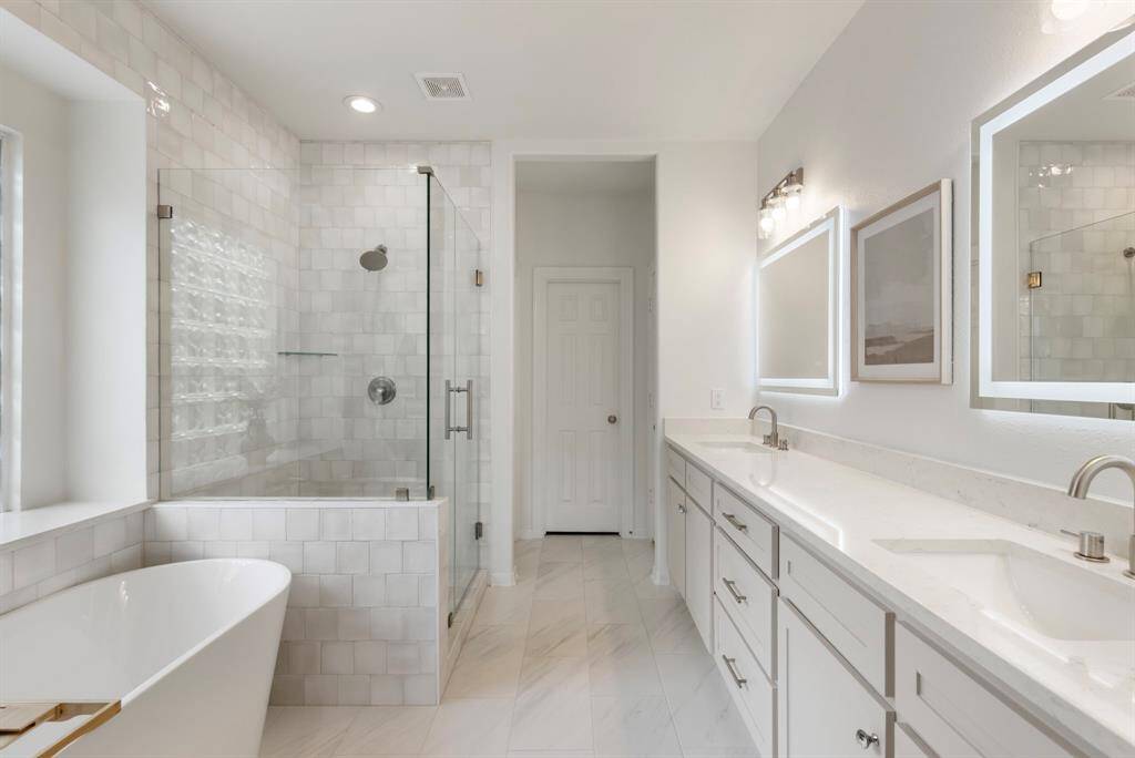 This bathroom features a modern, clean design with a spacious layout. It includes a freestanding bathtub, a glass-enclosed shower, dual sinks with a long countertop, and elegant Halo lighted mirrors. The neutral color palette enhances the bright and airy feel.