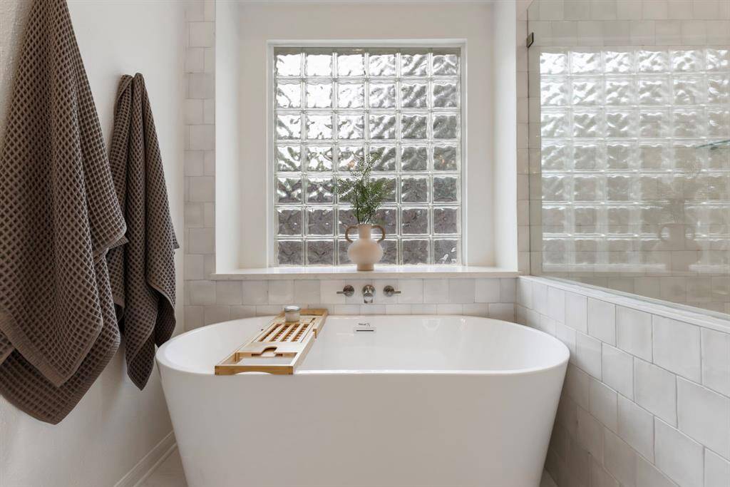 This bathroom features a modern, freestanding tub with a wooden tray, set against a wall of glass blocks that allow natural light. It includes a sleek tiled shower area creating a serene, spa-like atmosphere.
