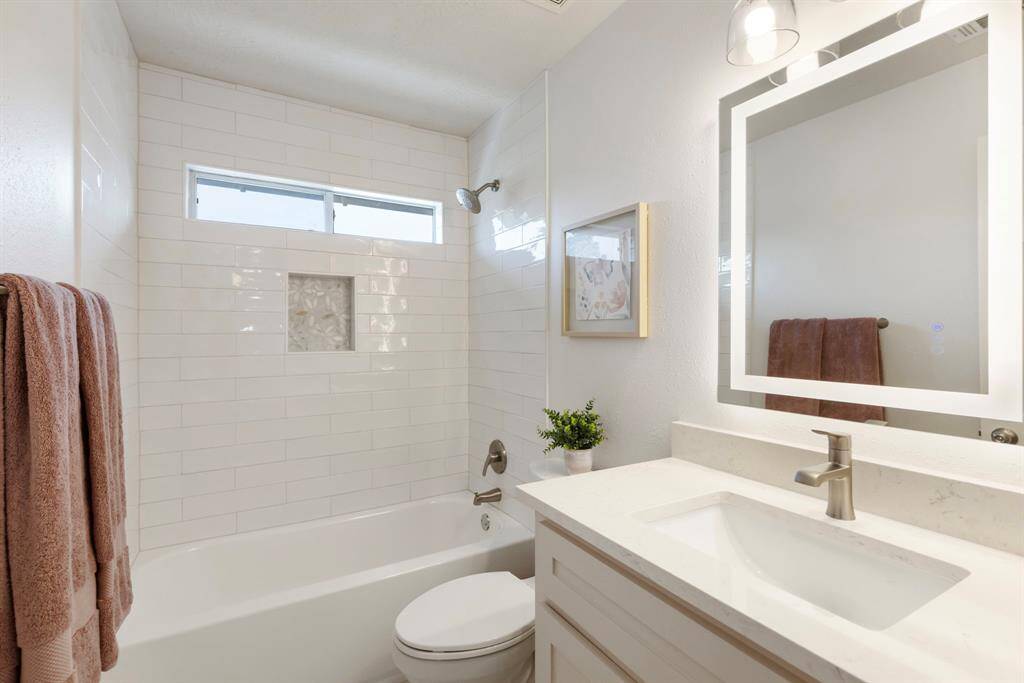 This bathroom features a clean, modern design with white subway tiles, a built-in tub, and a large illuminated Halo mirror. It has stylish fixtures, a small window for natural light, and tasteful decor, creating a bright and inviting space.