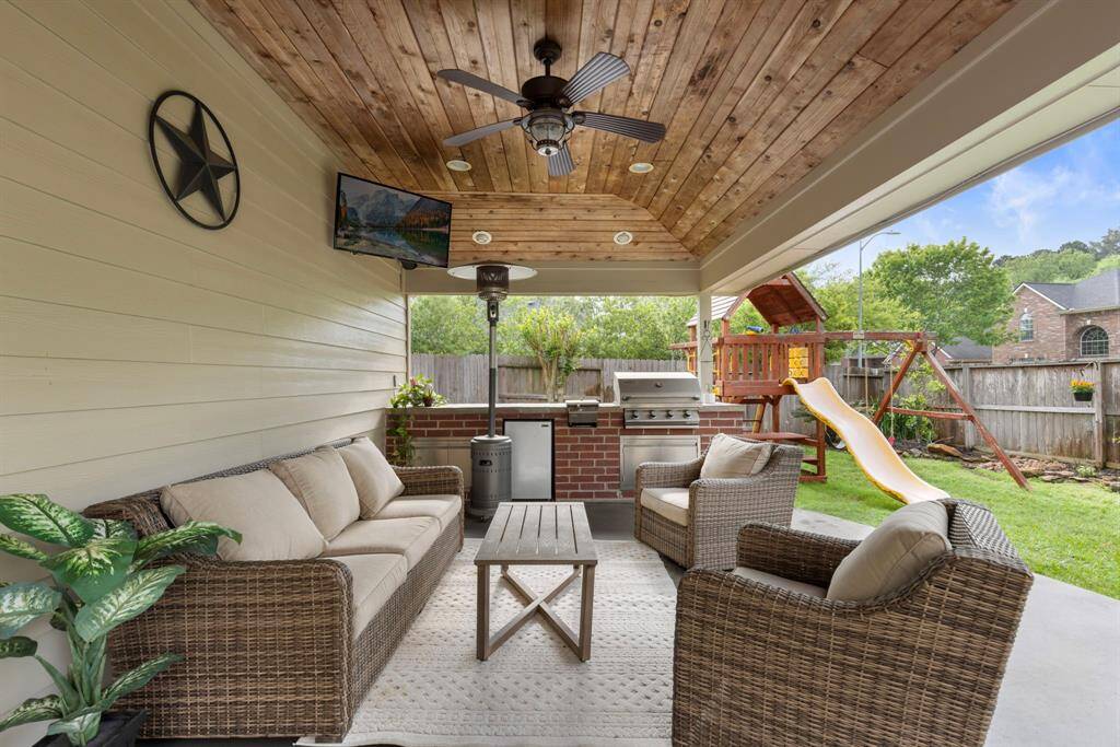 This outdoor living space features a covered patio with seating area, a ceiling fan, and a mounted TV. There's a built-in grill and countertop for entertaining, and a play set with a slide in the fenced backyard. Perfect for family gatherings.