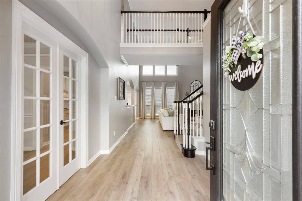 This inviting entryway features a glass-paneled front door, light wood floors, and bright walls. French doors lead to an adjacent room, and a staircase with white balusters adds elegance. Large windows at the back bring in natural light, creating an airy, open feel.