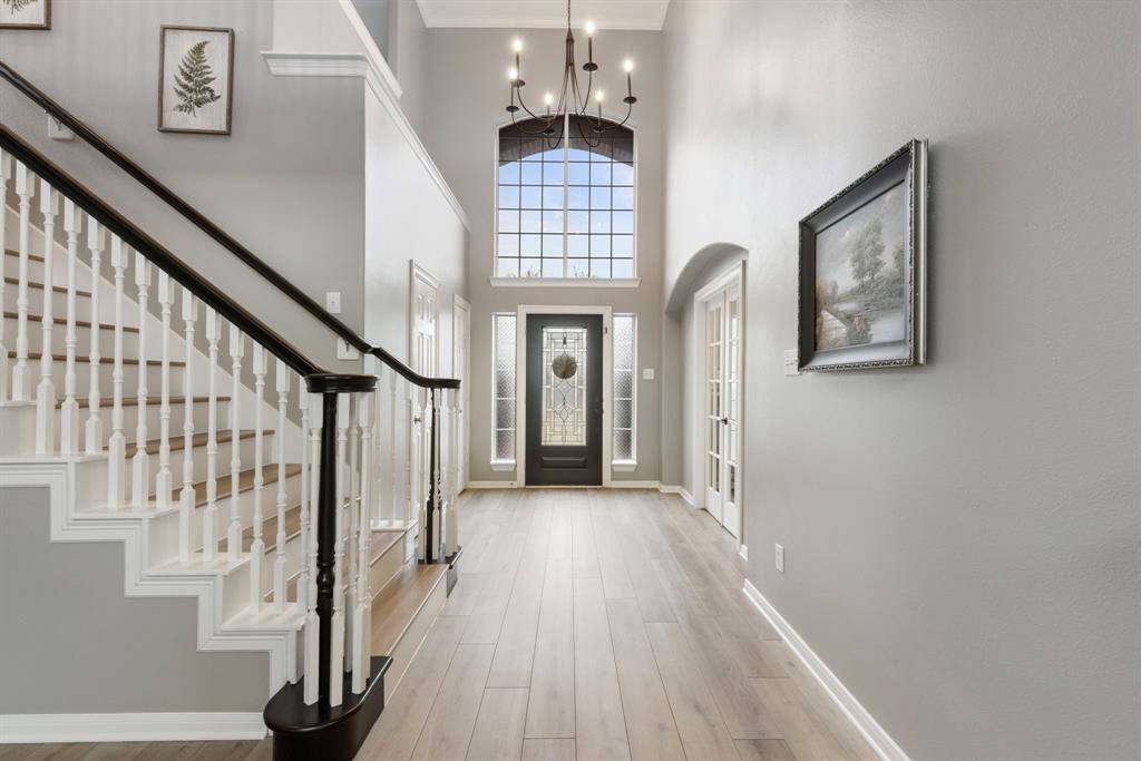 This spacious entryway features a high ceiling with a modern chandelier, a large window for natural light, and elegant wood flooring. The staircase has a classic design with white spindles and a dark handrail. French doors and tasteful wall art add a sophisticated touch.