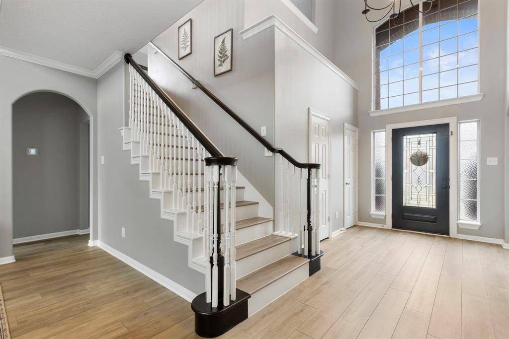 This photo showcases a bright, welcoming entryway with high ceilings and large windows. It features a stylish staircase with dark wood handrails, light wood flooring, and a decorative glass front door, creating an inviting atmosphere.
