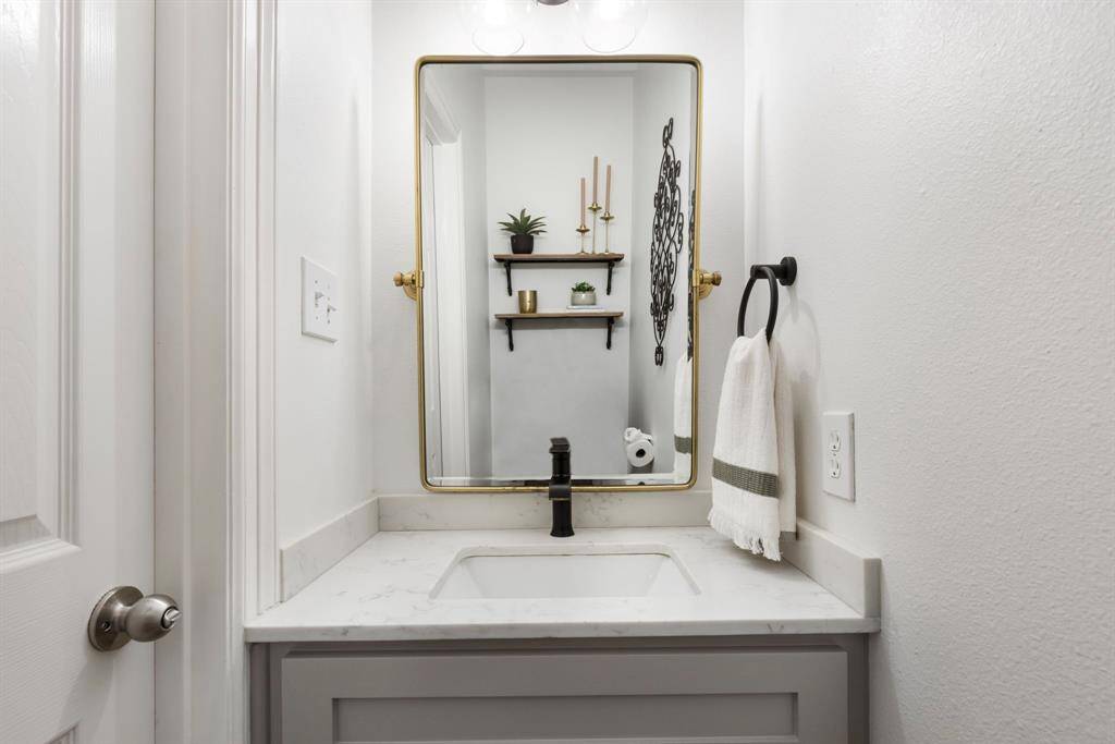 This is a modern, compact bathroom featuring a sleek vanity with a marble countertop, a stylish black faucet, and a rectangular mirror with a gold frame. The space includes elegant wall decor and shelves for added character and functionality.