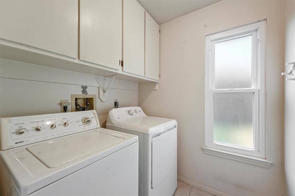 A bright and functional laundry room featuring a washer, dryer, overhead storage cabinets, and a frosted window for natural light. The washing machine and dryer stay with the home.