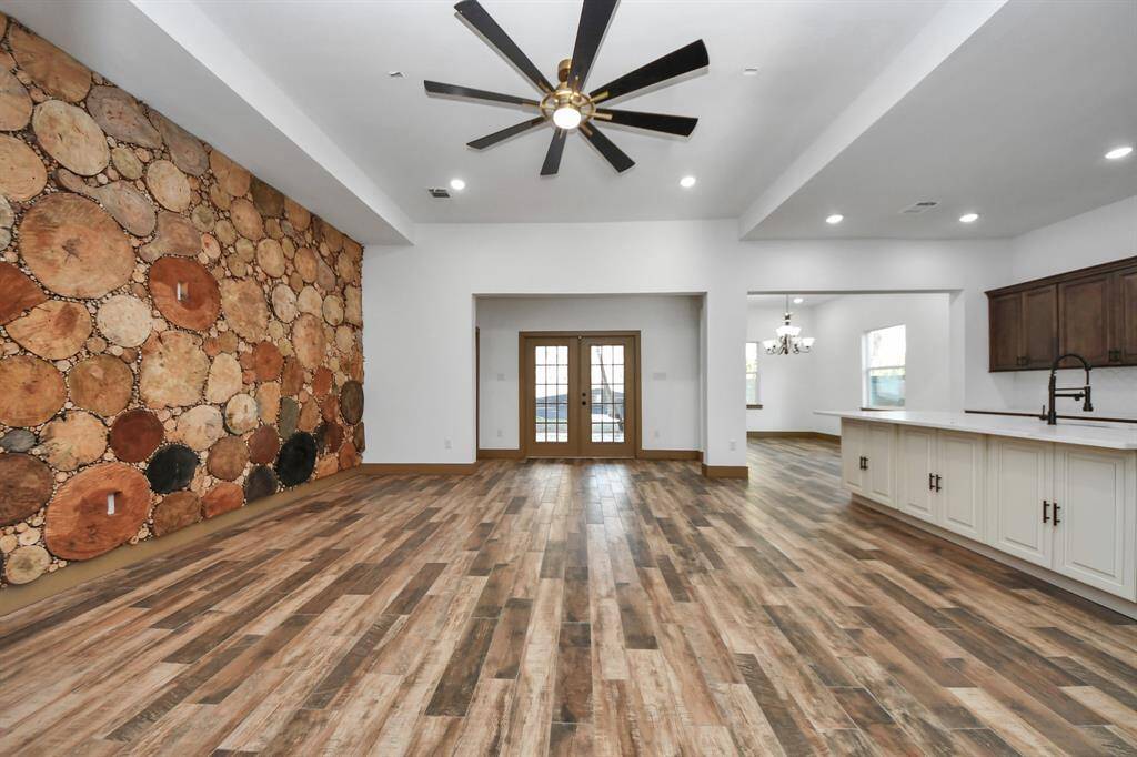 This spacious open-concept living area features a unique wood accent wall and stylish tile wood-look flooring. The room is bright with recessed lighting and a large ceiling fan, leading to a modern kitchen with ample cabinetry and a large island, perfect for entertaining.