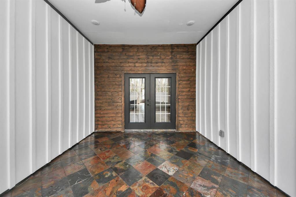 Outdoor patio includes a rustic wooden accent wall with double glass doors, flanked by sleek white paneling. The floor showcases a striking tile pattern and ceiling fan