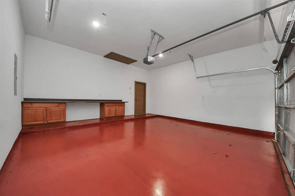 garage features a clean, red floor and white walls, with built-in wooden cabinets and a countertop for storage and workspace. It has an automatic garage door opener and ample lighting.