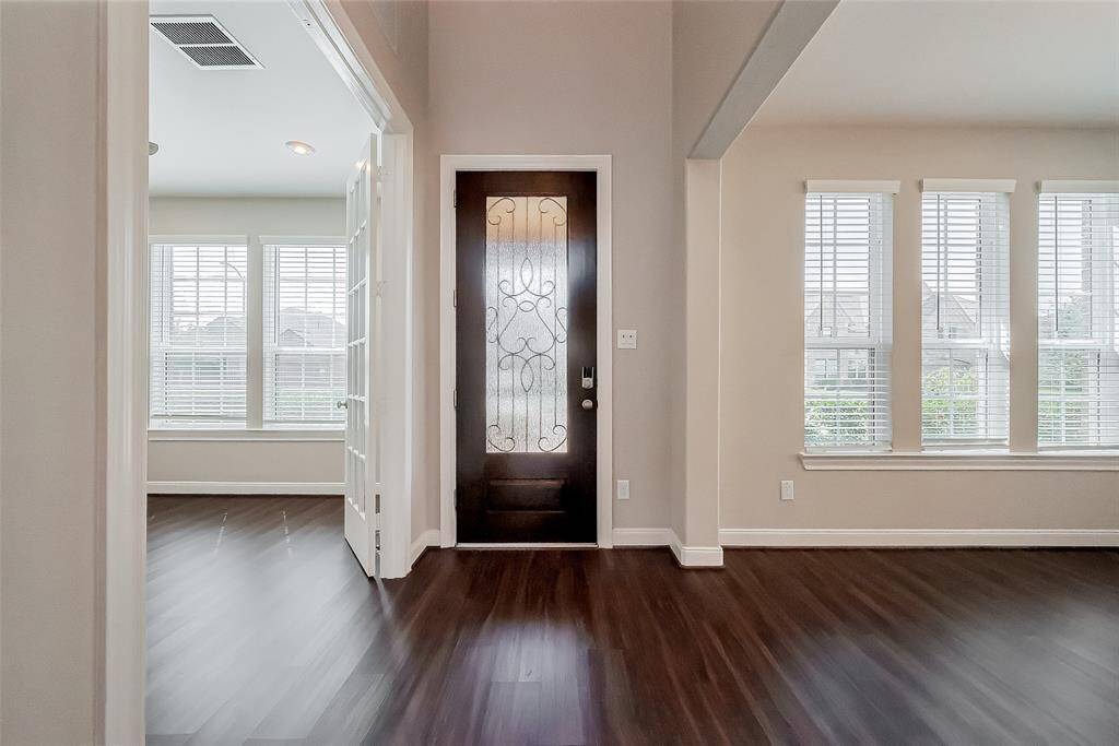 The glass-paneled front door, flanked by large windows, floods the entry with daylight, setting the stage for the inviting interior beyond. Dark wood-like laminate flooring adds contrast and elegance, creating a welcoming first impression.