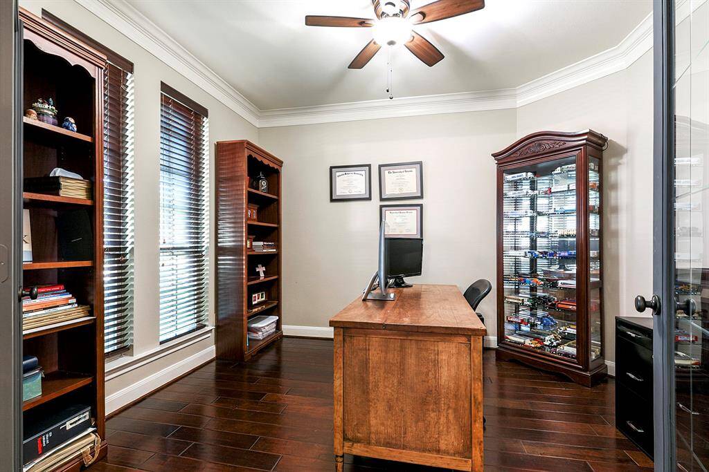 Step into the spacious home office and take note of the rich hardwood floors, elegant crown molding, and soaring ceilings. The room is complete with custom window treatments and a ceiling fan, blending comfort and style for the perfect work-from-home space.