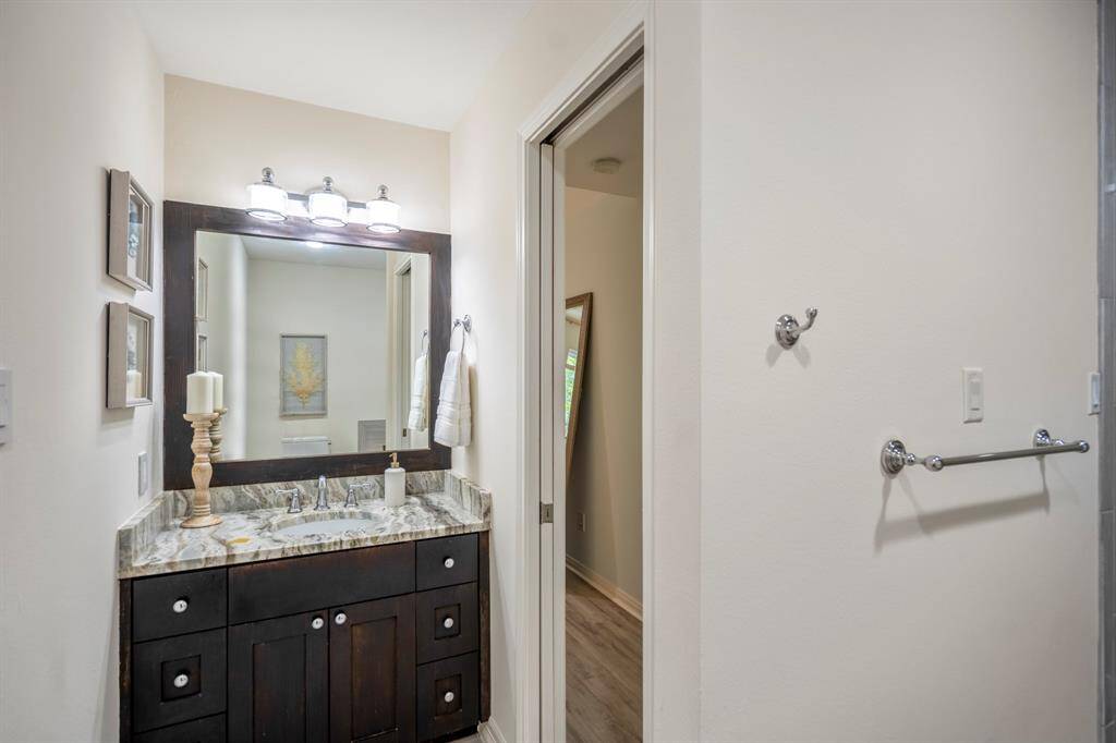 This bathroom also features a stylish dark wood vanity with a granite countertop, a large framed mirror with modern lighting above, and neutral wall colors.