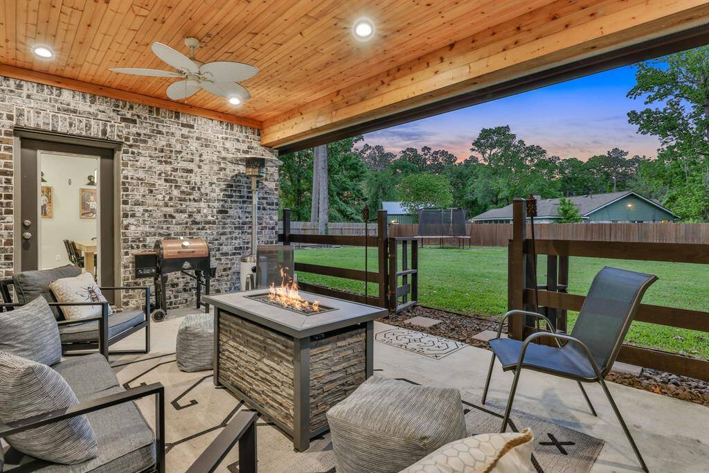 This inviting outdoor patio features a cozy seating area with a fire pit table, under a warm wood-paneled ceiling with ceiling fans. The brick walls add charm, while the mounted TV provides entertainment for gatherings.