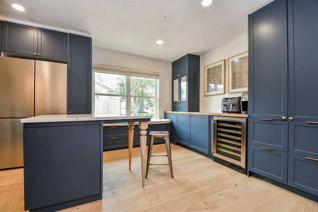 This kitchen features sleek, dark blue cabinetry with gold hardware, a modern stainless steel refrigerator, and a wine cooler. The wooden floors and large window provide a bright, inviting atmosphere, while a small island offers extra workspace.