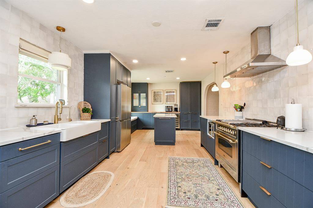 This kitchen features a modern design with sleek navy cabinetry and brass hardware. It includes stainless steel appliances, a farmhouse sink, and a central island. Light wood flooring and ample natural light create a warm, inviting atmosphere.