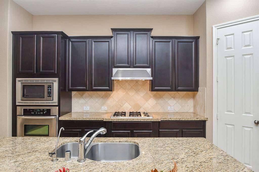 Modern kitchen with dark wood cabinets, stainless steel appliances, and granite countertops. Features a built-in oven, microwave, gas stove, and double sink. Light tile backsplash complements the elegant design.