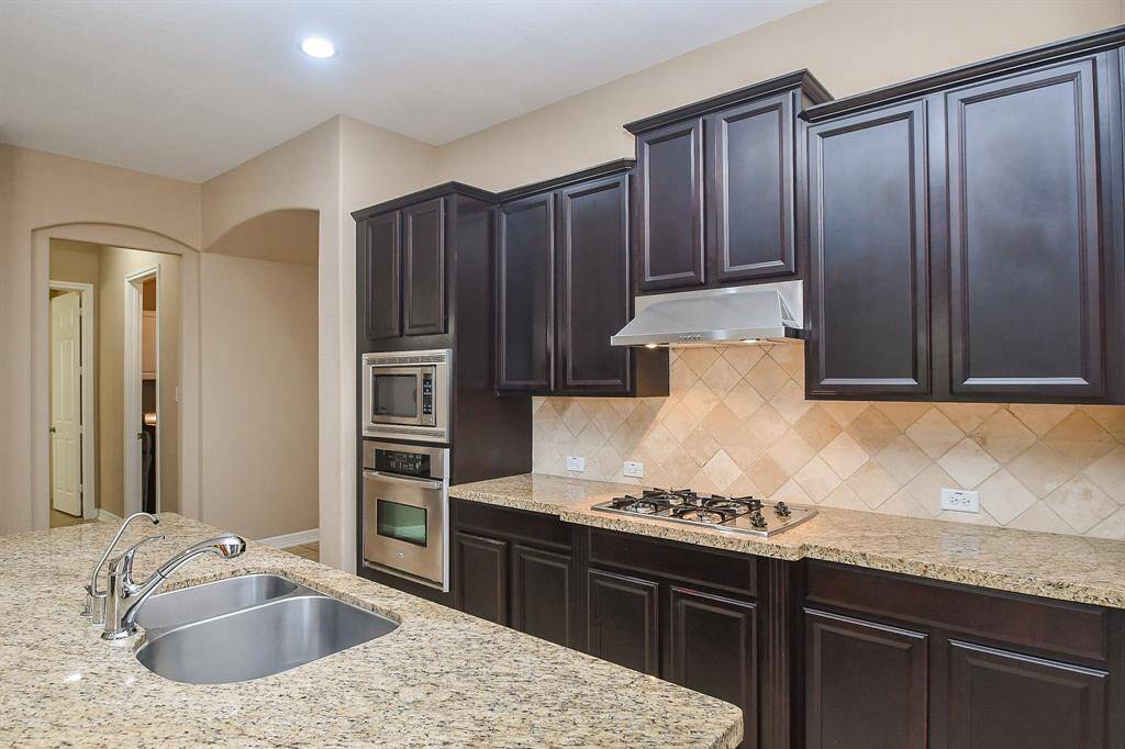This kitchen features elegant dark wood cabinetry, granite countertops, and a modern stainless steel cooktop and hood. It includes double ovens, a built-in microwave, and a spacious island with a double sink, perfect for meal prep and entertaining.