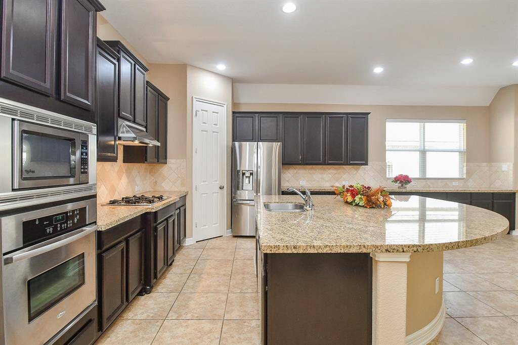This kitchen features dark wood cabinetry, granite countertops, and stainless steel appliances. It includes a built-in oven, a gas cooktop, a large island with a sink, and ample natural light from a wide window.