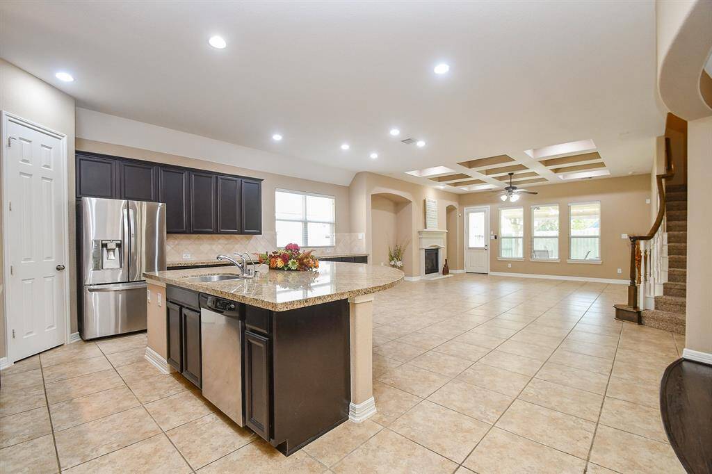 This spacious open-concept living area features a modern kitchen with granite countertops, stainless steel appliances, and dark cabinetry. The room is well-lit with recessed lighting and has large windows, a cozy fireplace, and tile flooring. A staircase leads to the upper floor, adding to the home's elegant design.