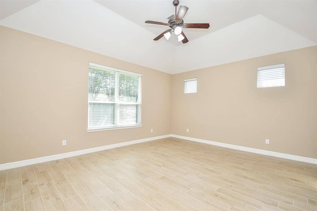 This spacious room features light wood-style flooring and neutral walls, with ample natural light from a large window and two smaller ones. A ceiling fan adds comfort and style.