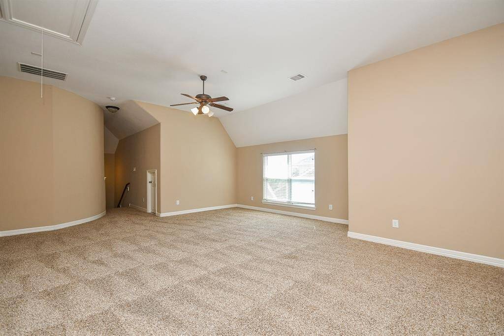 Spacious carpeted room with neutral walls, angled ceiling, and a ceiling fan. Features a large window for natural light and a hallway leading to other areas. Ideal for a bedroom or living space.