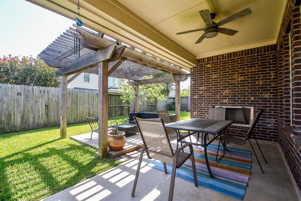 This photo showcases a cozy backyard patio featuring a wooden pergola, a ceiling fan, and a brick wall with a fireplace. The space includes outdoor dining furniture and a colorful rug, ideal for relaxing and entertaining.