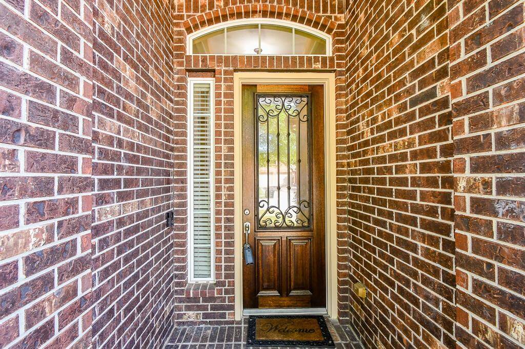 This photo showcases a welcoming entryway with a stylish wooden door featuring ornate ironwork, framed by red brick walls. A transom window above and a side window with blinds add natural light, complementing the inviting "Welcome" doormat.
