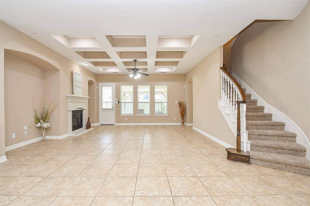 This spacious living area features a tiled floor, coffered ceiling, and a cozy fireplace. Natural light fills the room through large windows, and a carpeted staircase leads to the upper level. The neutral color palette creates a welcoming atmosphere.