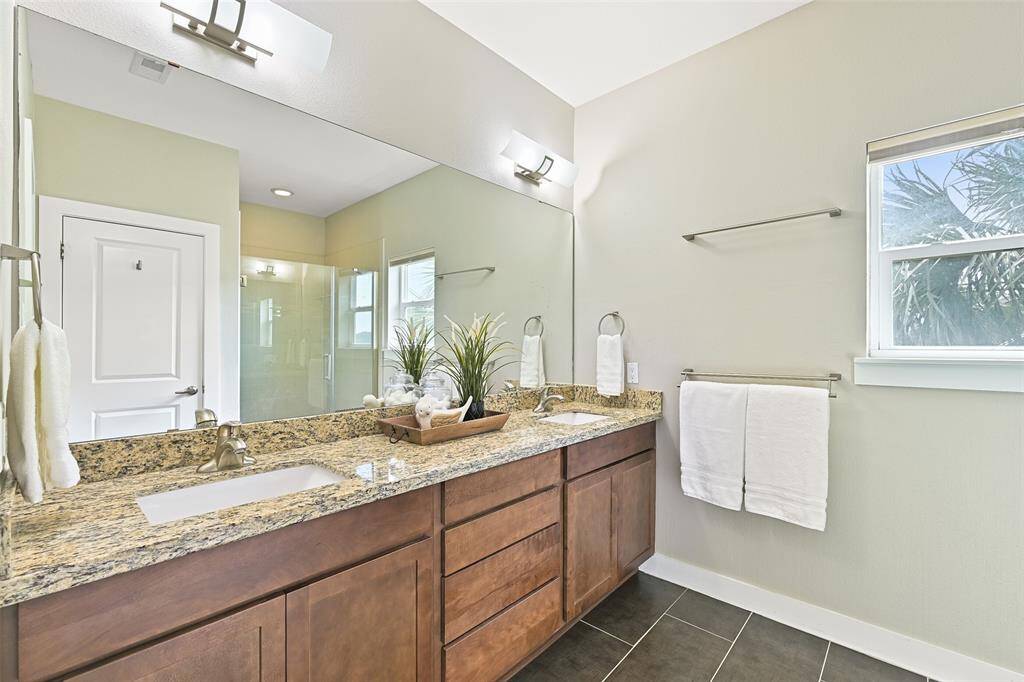 In the en suite master bathroom, find a lengthy vanity with gleaming granite countertops, dual rectangular basin sinks with brushed nickel faucets, stained cabinetry, tile floors, a large mirror, and contemporary lighting.