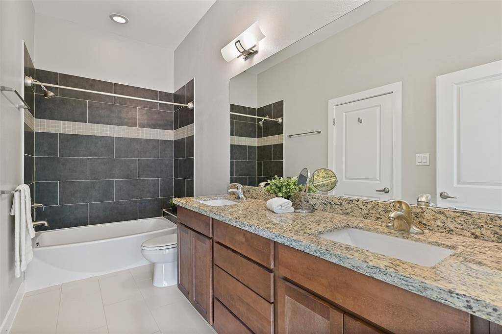 This bathroom is accessible from both the previous bedroom and the main living area. Here, you'll find a lengthy granite countertop with dual rectangular basin sinks, stained wood cabinetry, tile floors, contemporary lighting, and a combination bathtub and shower with a modern tile surround.