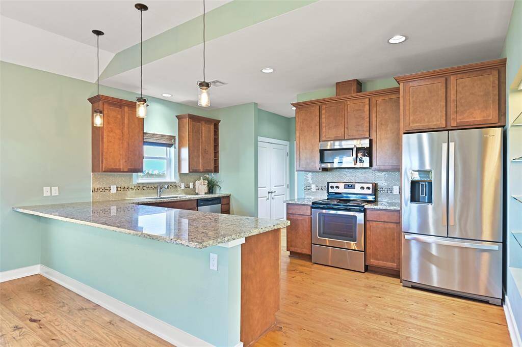 A well-appointed peninsula-style kitchen features gleaming granite countertops with enough space for 4-5 barstools that create additional seating. Also note the simple & contemporary pendant lighting and the glass tile backsplash.