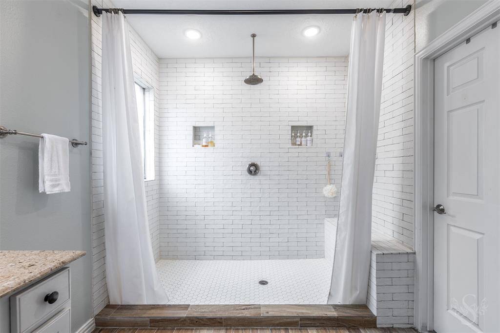 private ensuite that has been remodeled with a massive walk-in shower with bench seat, overhead rain shower head, freshly painted cabinets, and wood look tiled flooring.