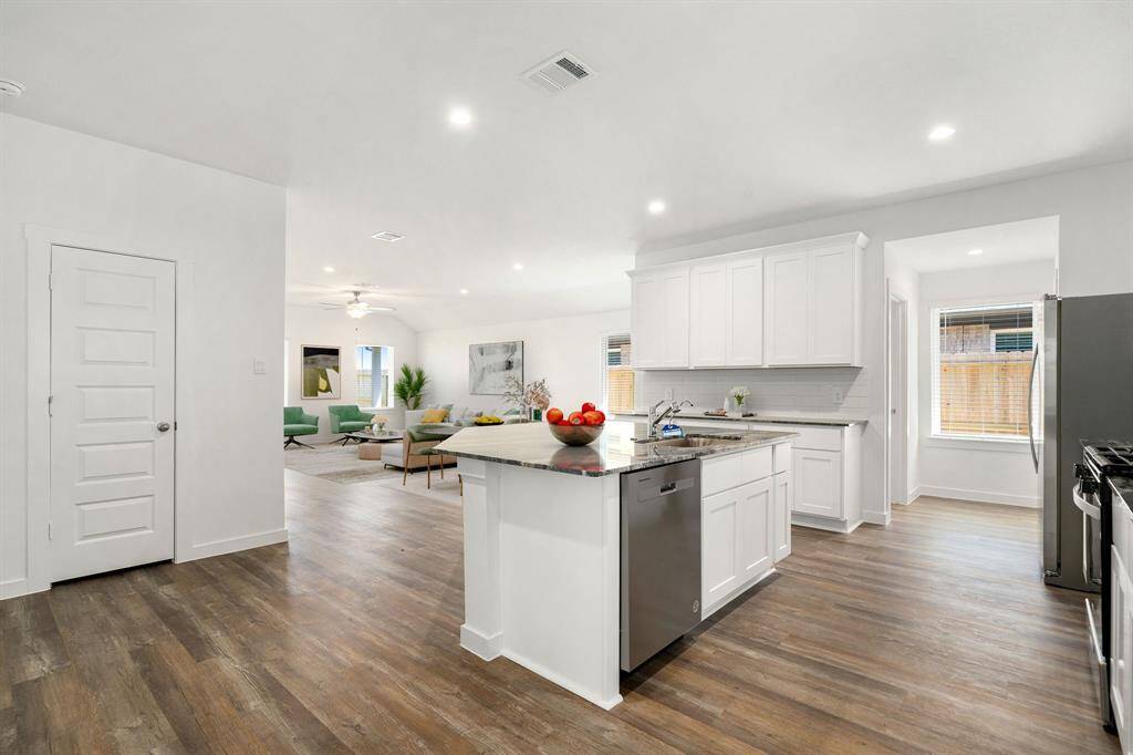 This bright and spacious kitchen features sleek white cabinetry, granite countertops, a large center island with sink, and modern stainless steel appliances. The open-concept layout flows seamlessly into the living area, enhanced by recessed lighting and durable vinyl plank flooring throughout. Ample natural light pours in from large windows, creating a warm and inviting atmosphere perfect for everyday living and entertaining.