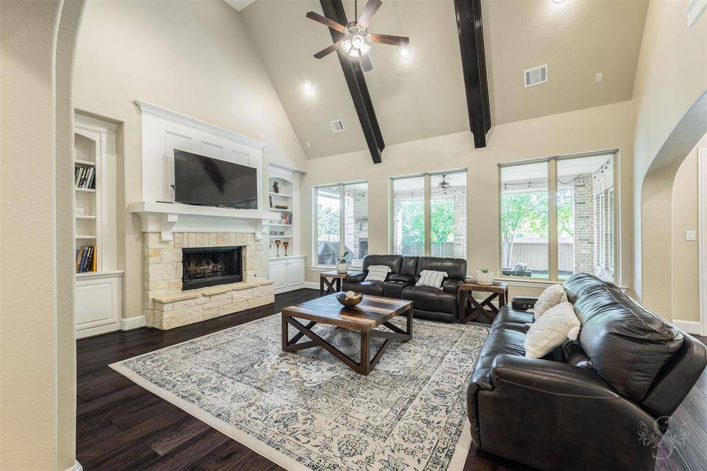 This spacious living room features high vaulted ceilings with dark wood beams, a cozy stone fireplace, built-in shelving, and ample natural light from large windows. It offers a welcoming, open-concept space perfect for relaxation and entertaining.