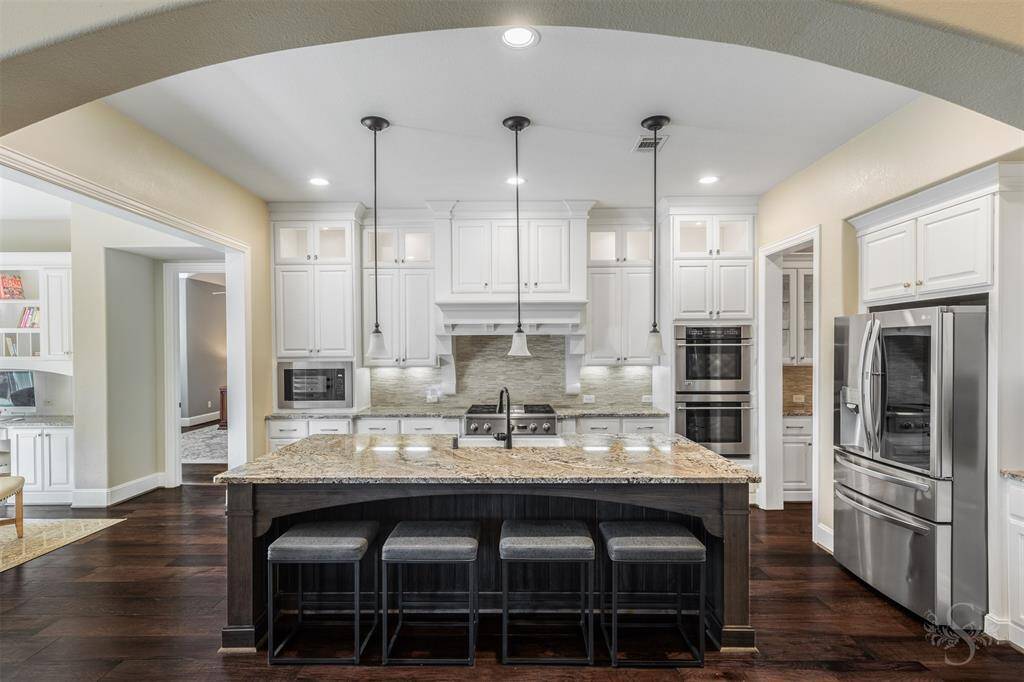 This modern kitchen features white cabinetry, granite countertops, and stainless steel appliances. A large island with pendant lighting and bar seating offers a perfect spot for casual dining. Rich hardwood floors add warmth and elegance, and the open layout flows seamlessly into adjacent living areas.