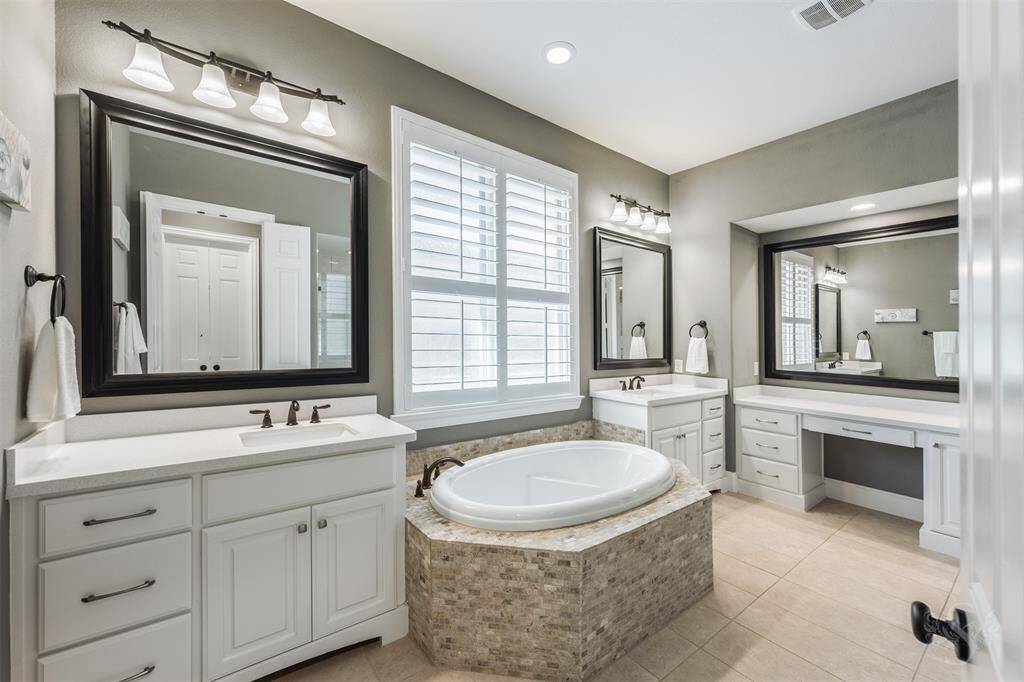 This spacious bathroom features a central soaking tub with a tiled surround, dual vanities with dark-framed mirrors, ample counter space, and built-in storage. Natural light streams in through a large window with shutters, creating a bright and inviting atmosphere.