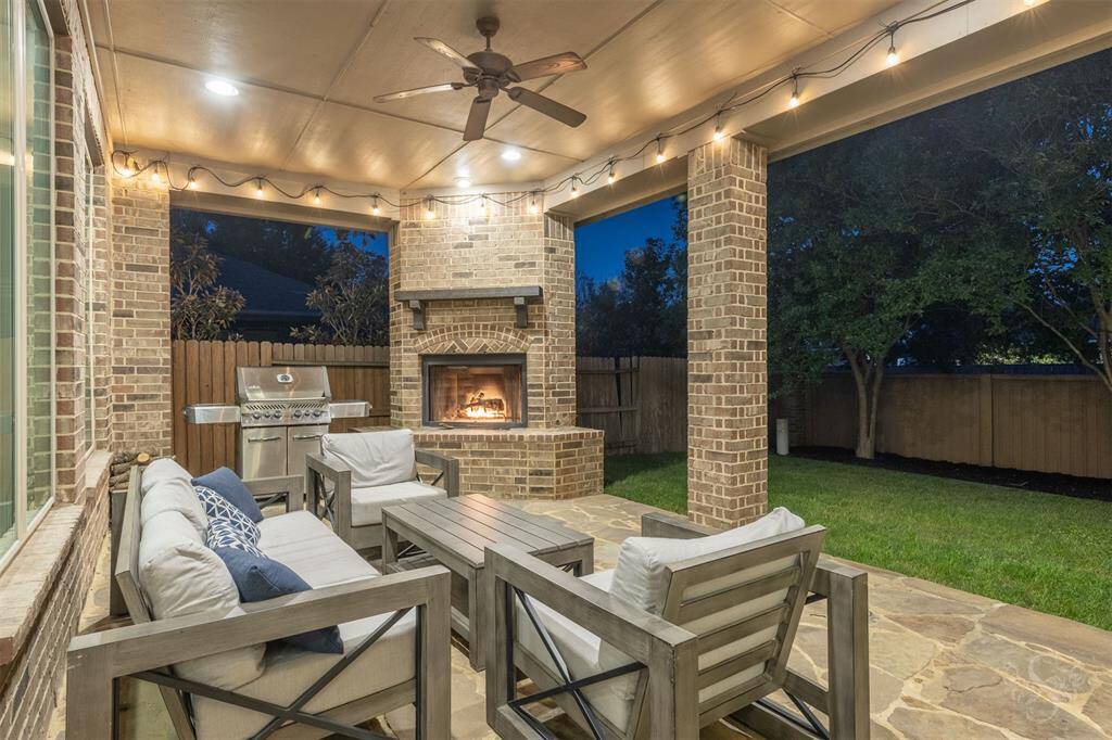 This inviting outdoor patio features a cozy seating area with cushioned chairs and a fireplace. It includes a built-in grill, string lights, and a ceiling fan, all under a covered, brick-accented space overlooking a lush lawn. Perfect for entertaining or relaxing evenings.