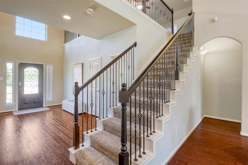 From this angle, the curved iron balusters and rich wood handrail highlight the craftsmanship of the staircase, while the soaring ceilings and front door sidelights fill the foyer with natural light—making a grand impression from the moment you walk in.