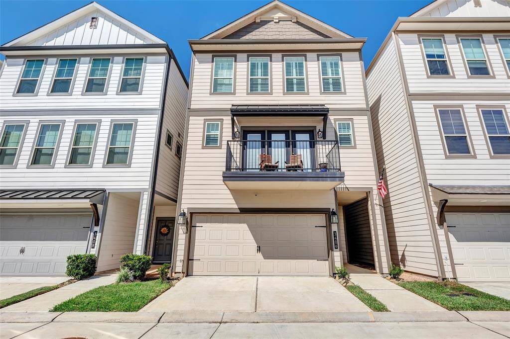 This is a modern, multi-story townhouse with a neutral color palette, featuring a garage, a small balcony on the second level, and a tidy front entrance. The architectural style is contemporary with clean lines.