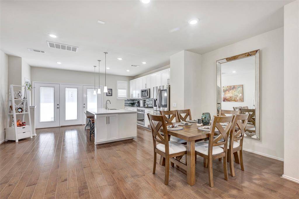 This is a bright, open-concept space featuring a modern kitchen with white cabinetry and an island, complemented by a spacious dining area with a large wooden table. The room is well-lit with recessed lighting and has easy access to the outdoors through double doors.