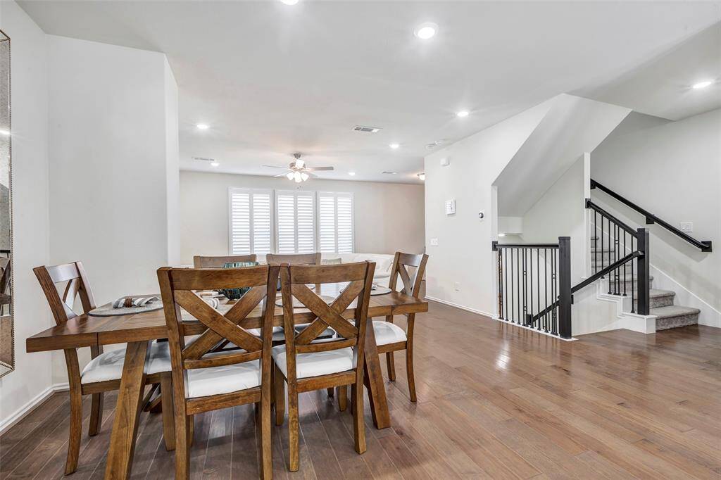 This is a spacious, modern dining area with hardwood floors, ample natural light from large windows, and a view of an elegant staircase leading to the upper level. The room is well-lit with recessed lighting and a ceiling fan.