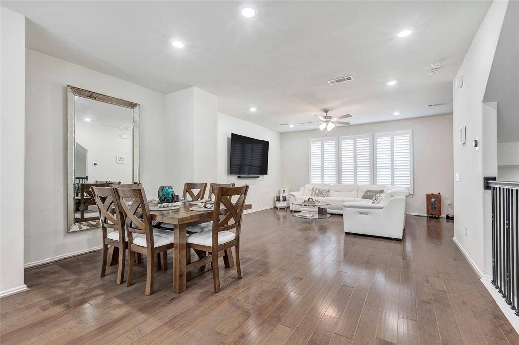 This is a spacious, well-lit open-concept living and dining area with wood flooring, recessed lighting, a large mirror accent, and plantation shutters. The space is modern and versatile, with a neutral color palette.