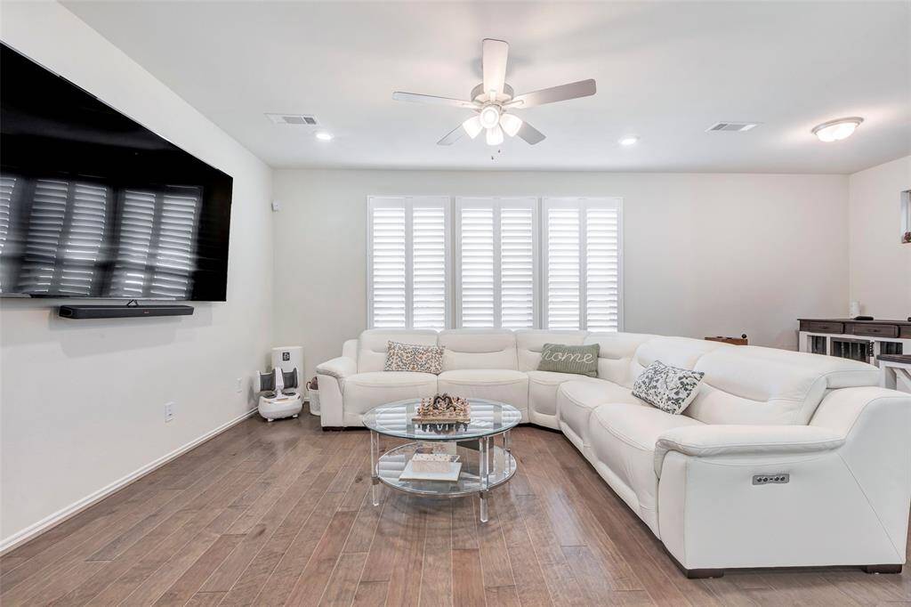 This is a bright, modern living room featuring hardwood floors, large windows with plantation shutters, and a neutral color palette. The space includes a mounted flat-screen TV and a comfortable white sectional sofa, with a cozy, inviting atmosphere.