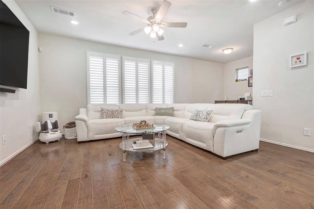 This is a well-lit, spacious living room featuring a large sectional sofa, hardwood floors, and plantation shutters on the windows. The room includes a ceiling fan and is pre-wired for a wall-mounted TV.