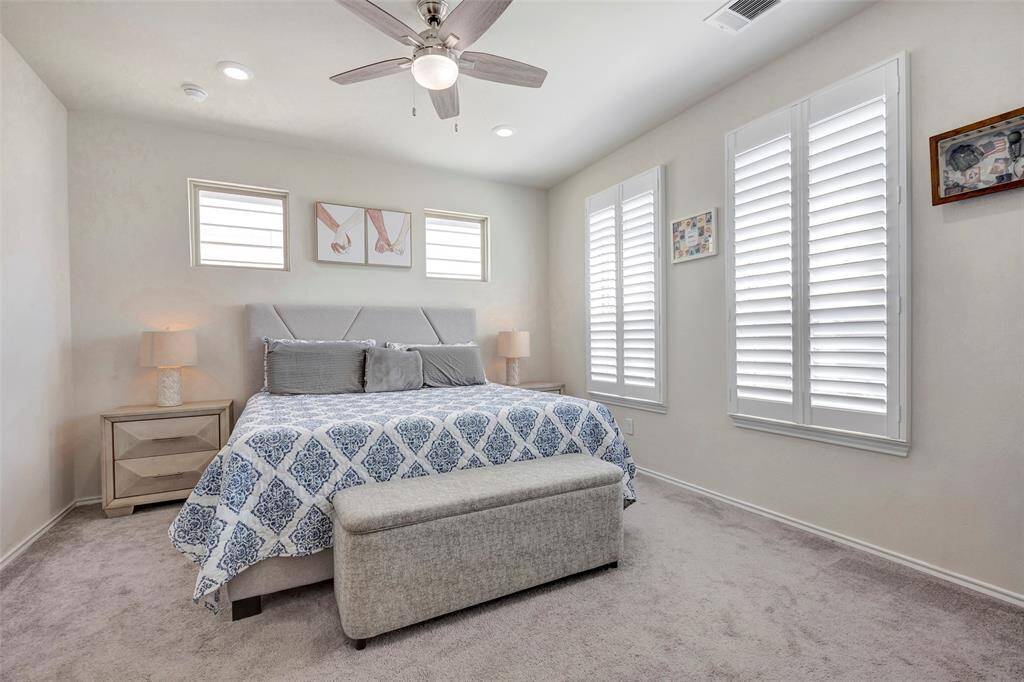 This is a well-lit, cozy bedroom featuring a large bed with an attractive blue and gray bedding set, two nightstands with lamps, a ceiling fan, plush carpeting, and three windows with plantation shutters offering ample natural light.