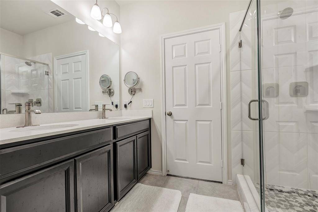 This is a modern, well-lit bathroom featuring a double vanity with dark cabinetry, a spacious glass-enclosed shower, and a neutral color scheme.