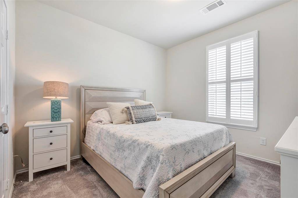This is a bright and airy bedroom featuring neutral wall colors, a large window with plantation shutters, and a modern bed with coordinating nightstand. The room is carpeted and has a clean, minimalist aesthetic.