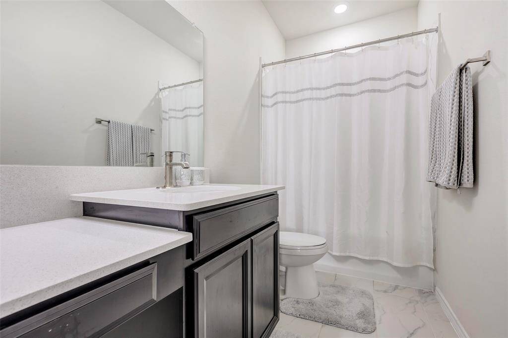 This is a modern, clean bathroom featuring a vanity with gray cabinetry and white countertop, a toilet, and a shower with a white curtain. The space is accented with neutral colors and simple, elegant decor.